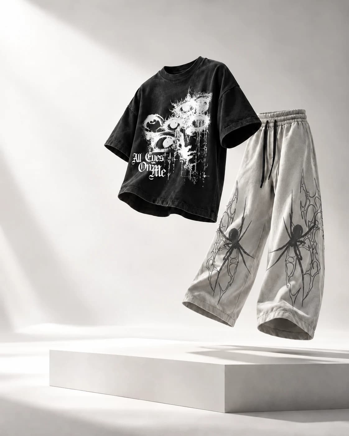 A floating black All Eyes On Me graphic tee and pale spider-print trousers over a white plinth